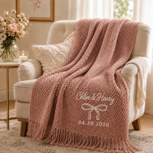 May include: A dusty rose knitted throw blanket with fringe edges, featuring white embroidered text that reads Chloe & Henry, a line-art bow graphic, and the date 04.30.2026, draped over a cream-coloured upholstered armchair with a matching pillow.