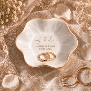 May include: A white ceramic ring dish with a scalloped edge and gold trim. The center features the gold text "j ♡ l", "Jessica & Lucas", and "12.06.2026". Two gold wedding bands rest inside the dish, while another ring sits on the textured fabric.