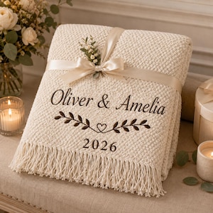 May include: A cream-colored knit throw blanket featuring a textured popcorn stitch pattern and long fringe at the bottom. The blanket is tied with a beige satin ribbon and embroidered with the text Oliver & Amelia, 2026, and a leaf motif.