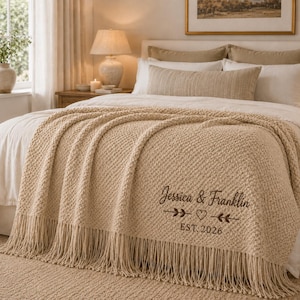 Könnte beinhalten: Eine beigefarbene Strickdecke mit Fransenbesatz, die mit dunkelbraunem Text bestickt ist, der Jessica & Franklin, ein Herzsymbol und den Text EST. 2026 zeigt. Die Decke hat ein strukturiertes Webmuster und liegt auf einem Bett.