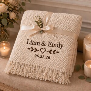 May include: A cream-colored textured throw blanket with fringe, tied with a satin ribbon and a small floral sprig. The blanket features dark brown embroidery reading Liam & Emily, a heart icon between two leafy branches, and the date 06.23.26.