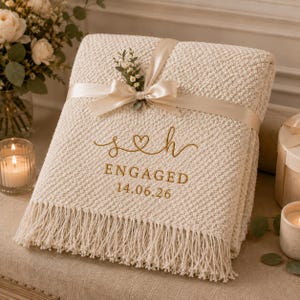 May include: A cream-coloured knitted throw blanket with fringe, tied with a satin ribbon and a small floral sprig. The blanket features gold embroidery reading s heart h, ENGAGED, and the date 14.06.26 centred on the front surface.