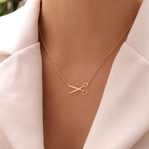 May include: A gold-coloured necklace with a scissors pendant. The scissors are a simple design, and the necklace is worn against a white top. The chain is delicate and the pendant is centered on the chest.