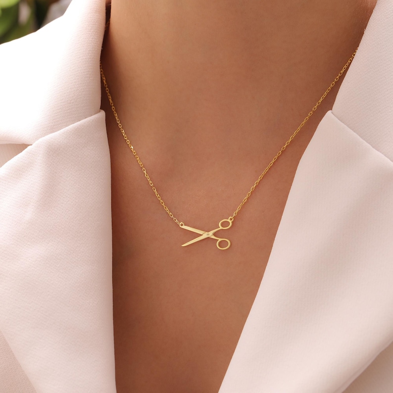 May include: A gold-coloured necklace with a scissors pendant. The scissors are a simple design, and the necklace is worn against a white top. The chain is delicate and the pendant is centered on the chest.