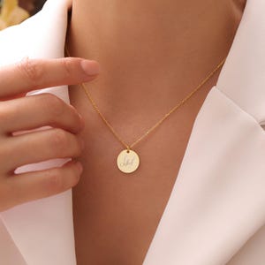 May include: A gold-colored necklace with a delicate chain and a round pendant. The pendant features a handwritten script. The necklace is worn against a white top.