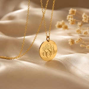 May include: A gold-toned necklace featuring a circular pendant with an engraved monogram of three interlocking letters. The pendant hangs from a delicate gold-toned chain, showcasing a polished, reflective surface that catches the light.