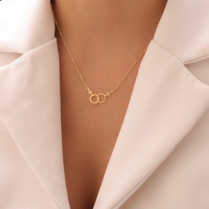 May include: A gold-colored necklace featuring two interlocking circles with gender symbols. The necklace is delicate and hangs on a chain. The background is a white garment.