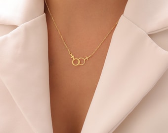 May include: A gold-toned necklace featuring a delicate chain and a pendant composed of two interlocking female gender symbols. The pendant is centered on the chain, which connects to the top of each circular symbol, creating a symmetrical design.