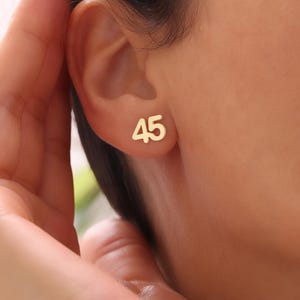 May include: Gold-colored stud earrings, each featuring the number "45". The earrings are displayed on an ear, showcasing their size and design. The earrings are a simple, yet elegant accessory.