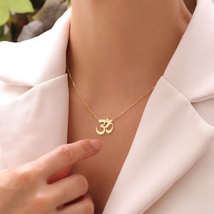 May include: A gold-colored necklace featuring the Om symbol pendant. The pendant is a stylized representation of the sacred syllable. The necklace is worn around the neck, and the chain is delicate. The background is a white blazer.