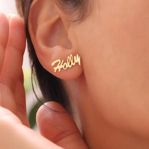 May include: Gold-colored stud earrings spelling the name "Holly" in a cursive script. The earrings are worn on an ear, with the name clearly visible. The design is simple and elegant, suitable for everyday wear.
