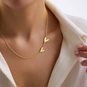 May include: A gold-colored necklace with two heart-shaped pendants. Each pendant has a name engraved on it. The necklace is worn around the neck, and the chain is delicate. The pendants are positioned to the side.