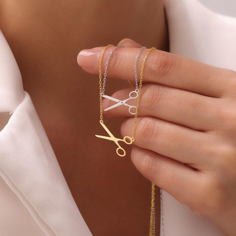May include: Two necklaces featuring scissor pendants. One necklace is gold-coloured, and the other is silver-coloured. The scissors are the central element of the necklaces, with a chain connecting to the scissors. The background is a soft, neutral colour.