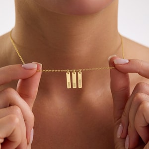 May include: A gold-colored necklace with three rectangular pendants. Each pendant is engraved with a name: "Avery", "Jade", and "Cadence". The necklace is held by a person.