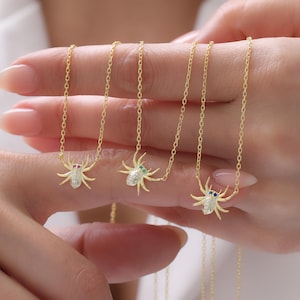 May include: Gold-toned spider pendant necklaces with gemstone accents. Each necklace features a detailed spider charm with a textured body and delicate legs. The spider's eyes are adorned with red, green, and blue gemstones, suspended from a fine chain.