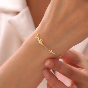 May include: A delicate gold bracelet featuring the name "Emilia" in cursive script, followed by a small cross charm. The bracelet is worn on a wrist, showcasing its elegant design and fine details. The bracelet is a beautiful piece of jewelry.