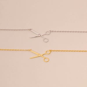 May include: Two necklaces featuring scissor pendants. One is silver, the other gold. The scissor charms are detailed and attached to fine chain necklaces. The background is a plain, light beige colour.