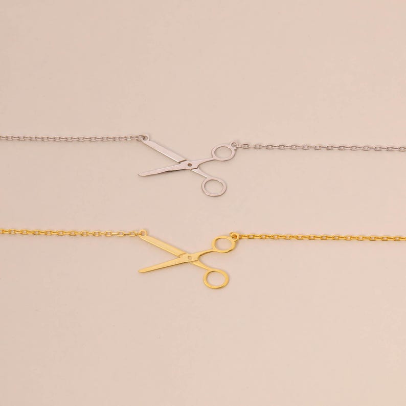 May include: Two necklaces featuring scissor pendants. One is silver, the other gold. The scissor charms are detailed and attached to fine chain necklaces. The background is a plain, light beige colour.