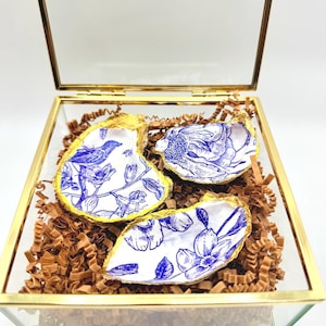 Könnte beinhalten: Eine Glas- und goldfarbene Box enthält drei dekorative Austernschalen. Die Schalen weisen blaue Blumen- und Vogelmuster mit goldfarbenen Rändern auf. Die Schalen liegen auf braunem Papier.