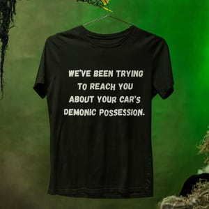 May include: Black t-shirt with white text: "WE'VE BEEN TRYING TO REACH YOU ABOUT YOUR CAR'S DEMONIC POSSESSION." The shirt is hanging on a metal hanger. A green monster hand with yellow claws is holding the hanger.