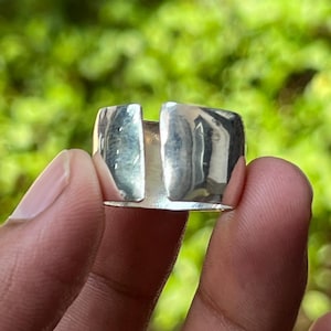 May include: A silver ring with a distinctive design. The ring has a thick band with a rectangular gap in the middle. The ring is held in a hand against a blurred green backdrop.