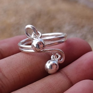 May include: A silver-colored ring with a spiral design. The ring features two small, round, dangling ornaments. The ring is shown on a hand against a blurred background.