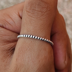 Puede incluir: Un anillo de plata con un diseño de cuentas. El anillo es delgado y presenta una serie de pequeñas cuentas espaciadas uniformemente alrededor de la banda. El anillo se usa en un dedo, mostrando su delicada y detallada artesanía.