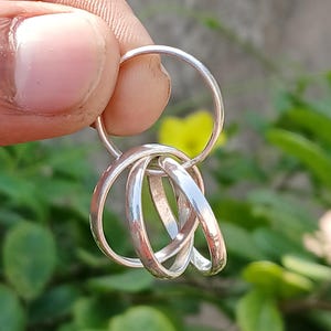 Op de afbeelding: Een zilverkleurige sleutelhanger met een grote ring en vier kleinere, in elkaar grijpende ringen. De ringen worden omhoog gehouden tegen een achtergrond van groene bladeren en een vage, neutrale achtergrond.