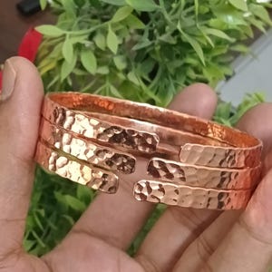 May include: Copper-colored hammered metal bracelet. The bracelet has a textured surface and an open design. The image shows the bracelet held in a hand, with green foliage in the background. The bracelet is a piece of jewelry.