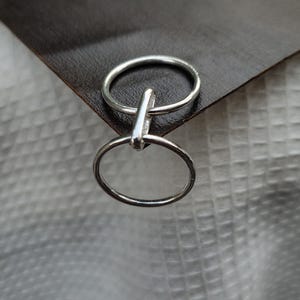 May include: Two silver rings connected by a small bar. The rings are circular and have a simple, minimalist design. The rings are displayed on a dark brown surface, with a white textured background.