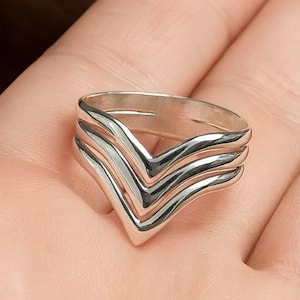 Może przedstawiać: Srebrny pierścionek z wzorem w kształcie szewronu. Pierścionek posiada trzy ułożone warstwowo paski w kształcie litery V, tworzące efekt warstwowy. Polerowany metal odbija światło, podkreślając geometryczny kształt pierścionka. Pierścionek jest pokazany na dłoni.