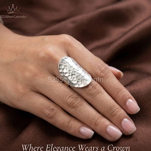 Puede incluir: Un anillo de plata grande y curvado con una textura martillada y con hoyuelos en toda su superficie. El anillo tiene grabado el texto 925SilverCrownIn y la frase Where Elegance Wears a Crown aparece en la parte inferior de la imagen.