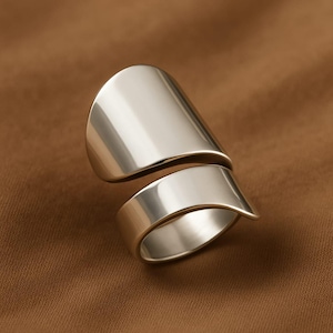 May include: A silver-colored ring with a modern design. The ring features a wide, curved band that wraps around the finger, with a second, overlapping band above. The polished metal reflects light, creating a sleek, minimalist aesthetic.