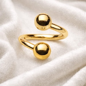 Puede incluir: Un anillo dorado con un diseño en espiral. El anillo presenta dos acentos esféricos en cada extremo de la espiral. El anillo se muestra sobre una superficie blanca y texturizada.