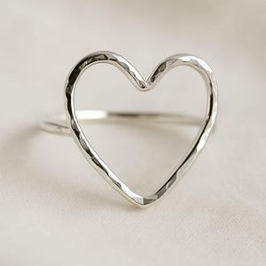 May include: A silver ring with a heart-shaped design. The ring features a hammered texture on the heart, creating a unique and eye-catching look. The band is thin and simple, complementing the heart shape. The ring is set against a soft, white background.
