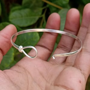 May include: A silver-colored bracelet with a knot design on one end and an open hook clasp on the other. The bracelet is held in a hand against a backdrop of green foliage. The bracelet's design is simple and elegant.