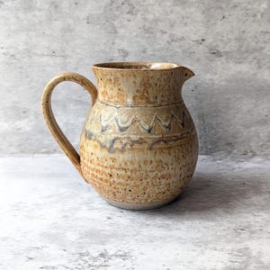 May include: A ceramic pitcher with a rounded body and a curved handle. The exterior features a speckled tan and beige glaze with a dark blue zigzag pattern encircling the upper section. The rim is slightly flared with a small pouring spout.
