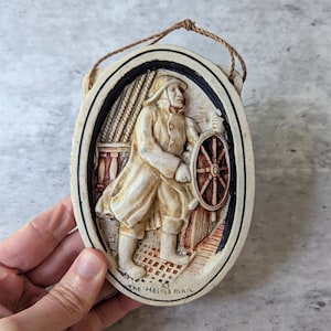 May include: An oval-shaped wall plaque featuring a relief sculpture of a sailor steering a ship with a wooden wheel. The figure wears a coat and hat, with the text The Helmsman inscribed at the bottom. The piece is cream-coloured with brown accents and includes a brown string for hanging.