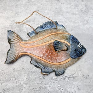 May include: A ceramic fish wall hanging featuring a mottled brown and blue glaze with smooth, raised orange curved lines across the body. The fish has a dark blue head with a single white eye and a jute twine loop attached for hanging purposes.