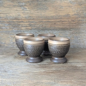 May include: Four ceramic egg cups with a textured, dark brown exterior and a lighter tan rim. The egg cups have a pedestal base and a smooth, light brown interior. The background is a wooden surface.