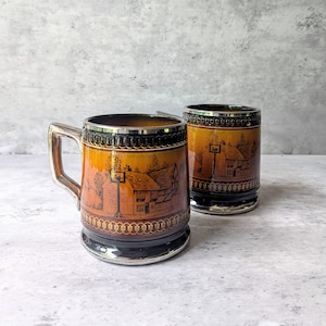 May include: Two ceramic mugs featuring a gradient amber and brown finish with black line art illustrations of a cottage and a signpost. Each mug has a metallic silver rim, a silver base, and a silver handle on the mug in the foreground.