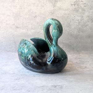 May include: A ceramic swan-shaped planter or decorative bowl featuring a glossy glaze with blended shades of deep black, dark teal, and vibrant turquoise. The swan has a curved neck and a hollowed-out body with a smooth, reflective finish.