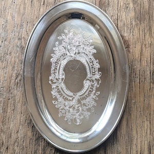 May include: Oval silver-plated serving tray with an elaborate floral and scroll pattern. The tray has a shiny finish and a decorative edge. The centre has an oval design with a dotted border. A vintage piece for serving or display.