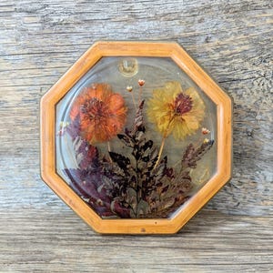 May include: An octagonal wooden frame encases dried flowers in resin. The arrangement features an orange and a yellow flower, with dark green and brown foliage. The frame is light brown and the background is a light grey.