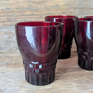 May include: Four vintage ruby red drinking glasses. The glasses have a textured, geometric pattern on the lower half and a smooth, rounded top. The glasses are made of a deep red glass and are arranged on a wooden surface.