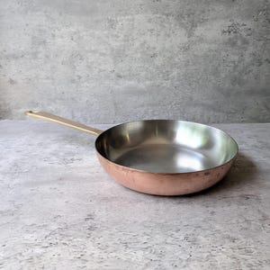 May include: A round copper frying pan with a polished silver-toned interior and a long, straight brass handle. The exterior of the pan features a warm, metallic copper finish with subtle variations in tone, while the handle is securely attached.