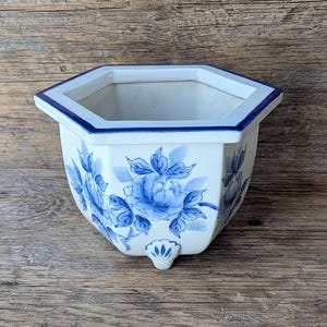 May include: White ceramic planter with a blue floral design. The hexagonal planter has a blue rim and a small foot. The blue floral pattern is hand-painted on the white surface. The planter is suitable for small plants.