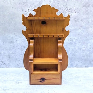 May include: A light brown wooden wall-mounted display rack featuring a decorative scalloped top edge, two horizontal shelves with notched grooves for holding items, a small open shelf section, and a single drawer with a visible wood grain knot.