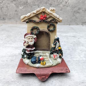 May include: A Christmas stocking holder featuring a miniature house with a central photo frame, a Santa Claus figure, a decorated tree, a snowman, and a red base. The house has a snow-dusted roof, a red bow, and two green wreaths hanging on the front.
