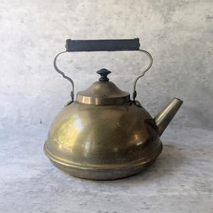 May include: A round brass tea kettle with a polished metallic finish, featuring a dark rectangular handle, a matching dark knob on the lid, and a short, angled spout. The kettle has a wide, curved body and a slightly domed lid with a central finial.